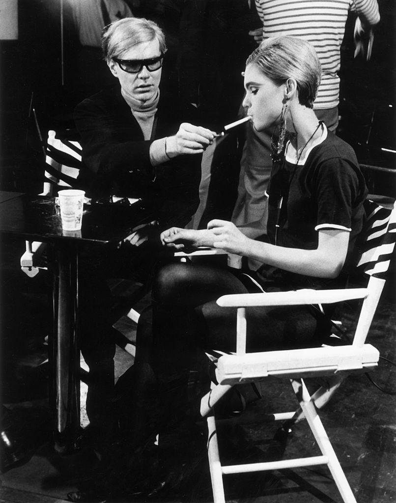 American Pop artist Andy Warhol (1928 - 1987) sits next to actor Edie Sedgwick (1943 - 1971) and lights her cigarette, on the set of one of his films